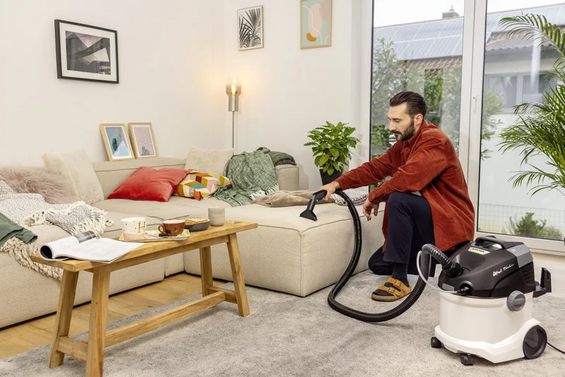 Person reinigt Sofa mit Kärcher Waschsauger in modernem Wohnzimmer. Tisch mit Tassen und Buch, Pflanzen und Bilder an der Wand.