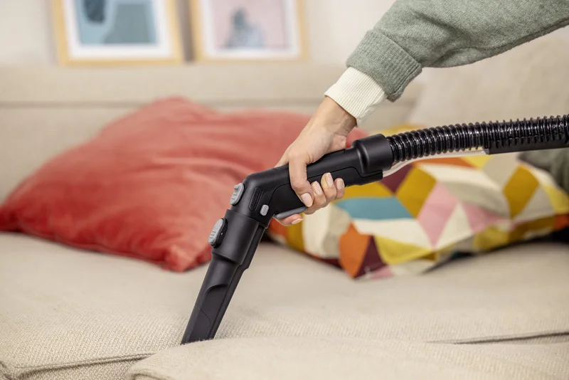 Hand hält Kärcher Staubsaugerrohr, reinigt beiges Sofa. Im Hintergrund bunte Kissen und gerahmte Bilder.