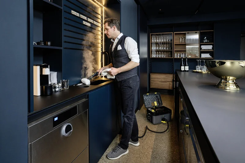 Mann reinigt Küchenspüle mit Kärcher Dampfreiniger in modernem Küchenraum. Wanddeko mit Schriftzug "Home Away From Home".
