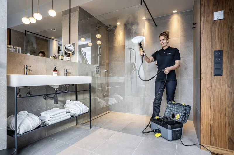 Person reinigt Duschkabine mit Kärcher Dampfreiniger in modernem Badezimmer. Waschbecken mit Handtüchern und Pflegeprodukten sichtbar.