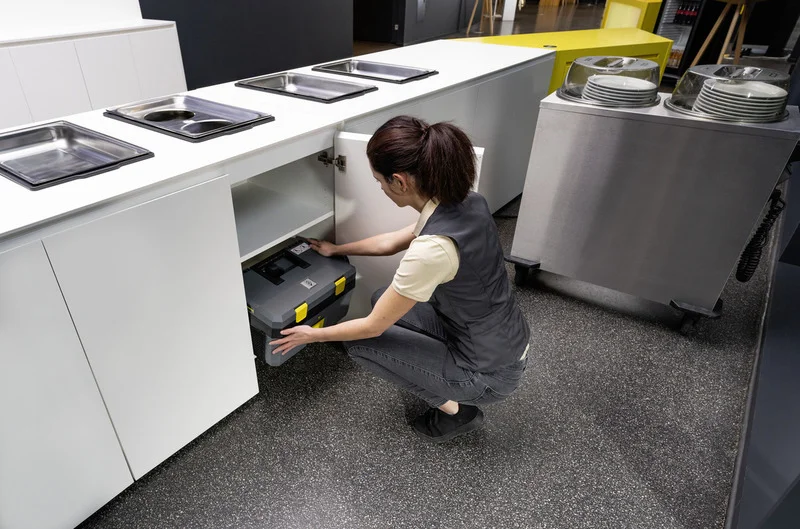 Person in Arbeitskleidung öffnet Küchenschrank und greift nach einem Kärcher Gerät. Umgebung mit Edelstahlmöbeln und Geschirrstapeln.