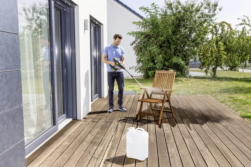 Mann reinigt Holzterrasse mit Kärcher Hochdruckreiniger, neben ihm ein Kanister. Terrasse vor einem modernen Haus, Garten im Hintergrund.