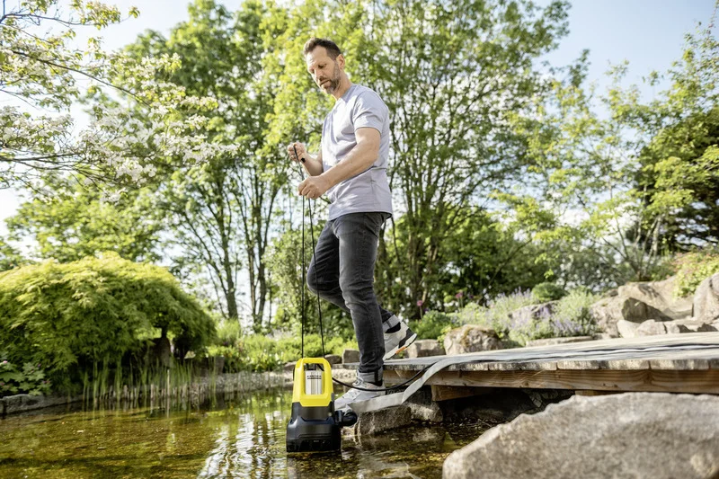 Mann steht auf Holzsteg über Teich und hält eine gelbe Kärcher Tauchpumpe, umgeben von grünen Bäumen und Pflanzen im Garten.