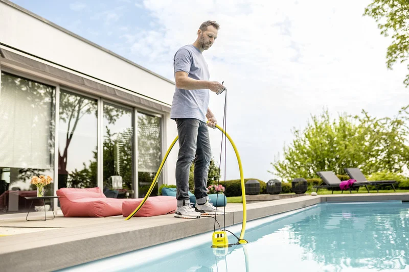 Mann steht am Pool und hält einen gelben Schlauch, der mit einer Kärcher Tauchpumpe verbunden ist. Im Hintergrund Gartenmöbel und Pflanzen.