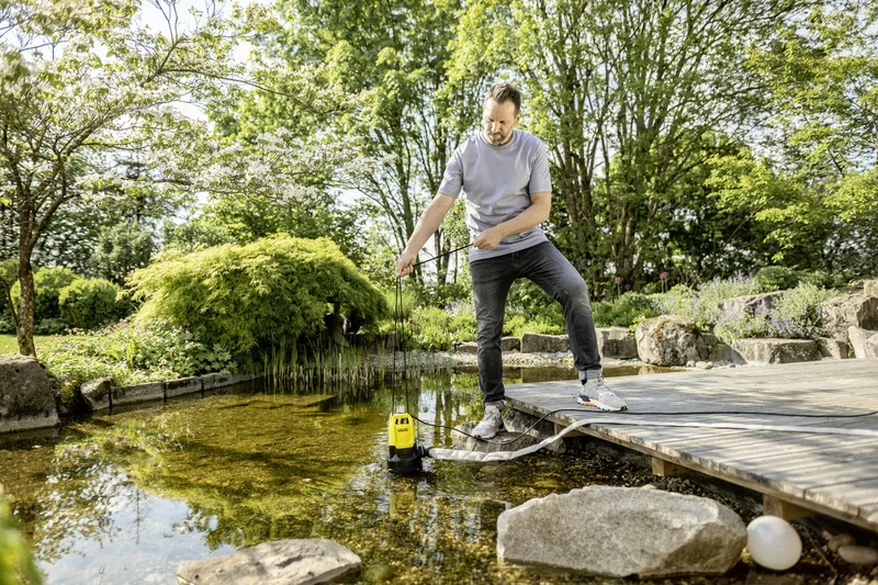 Mann steht auf Holzsteg und senkt eine Kärcher Tauchpumpe in einen Teich. Umgeben von üppiger Vegetation und Bäumen im Freien.