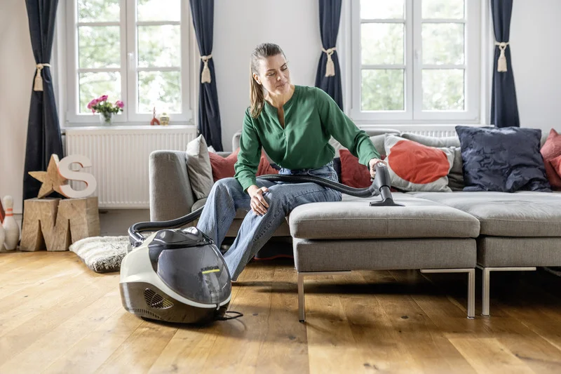 Person reinigt Sofa mit Kärcher Dampfreiniger in gemütlichem Wohnzimmer, Holzboden, Fenster mit Vorhängen, dekorative Kissen.