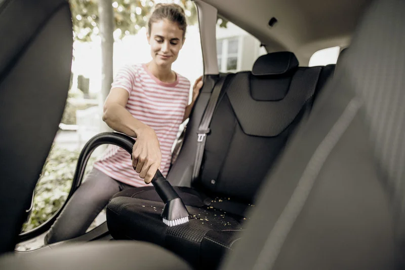 Person reinigt Autositze mit einem Staubsauger in einem Fahrzeug. Außenbereich mit Bäumen im Hintergrund.