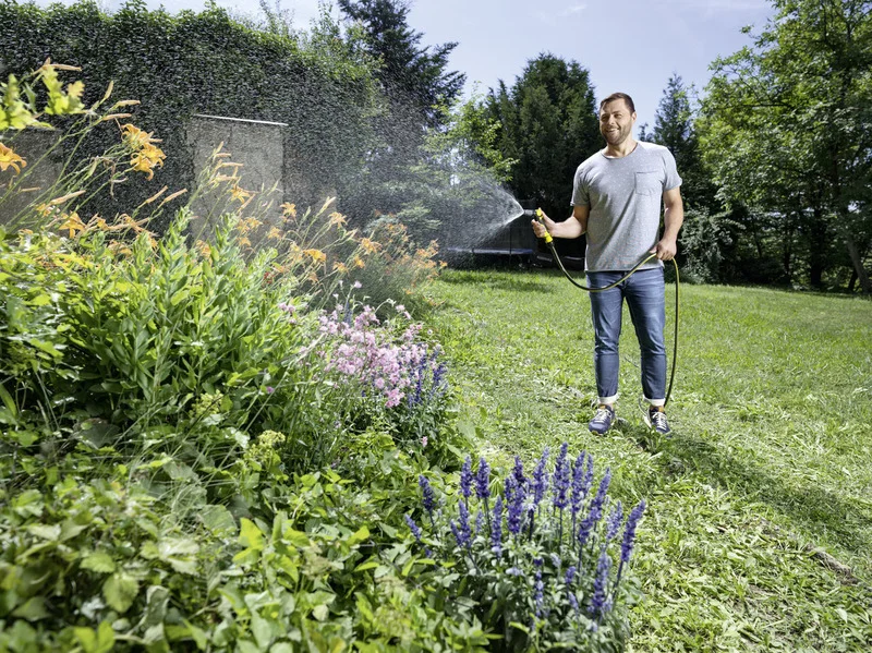 Person bewässert Blumenbeet im Garten mit einem Gartenschlauch. Im Hintergrund sind Bäume und ein blauer Himmel sichtbar.