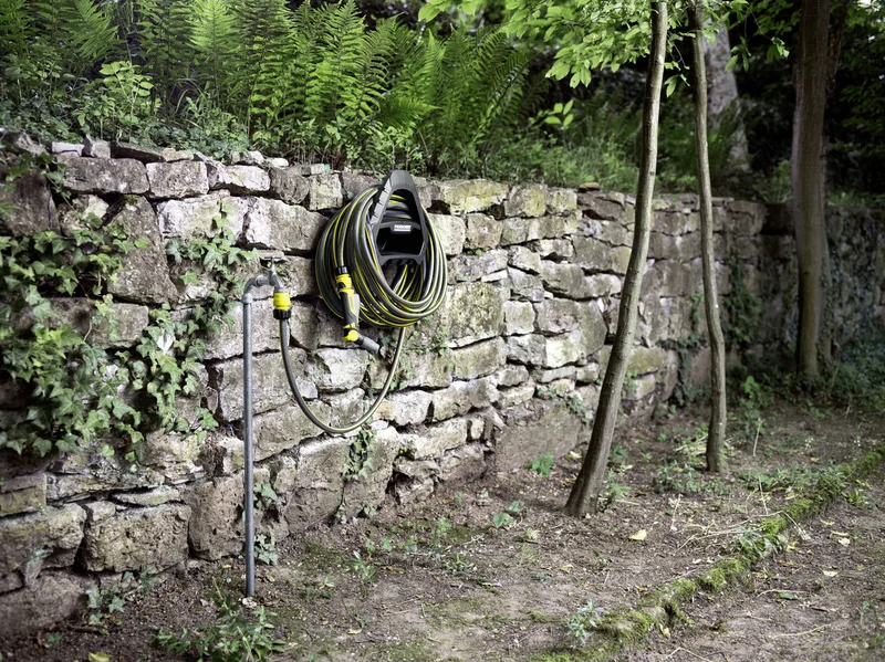 Gartenschlauch an Wandhalterung neben Wasserhahn, umgeben von Steinmauer und Pflanzen im Freien.