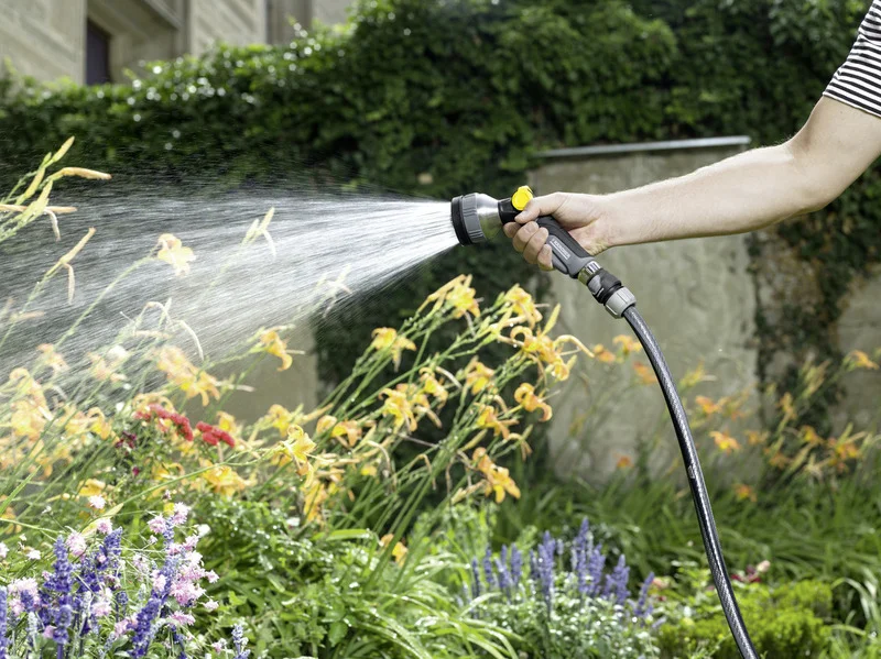 Person bewässert Gartenpflanzen mit Kärcher Gartenspritze. Im Hintergrund sind gelbe Blumen und grüne Hecke sichtbar.