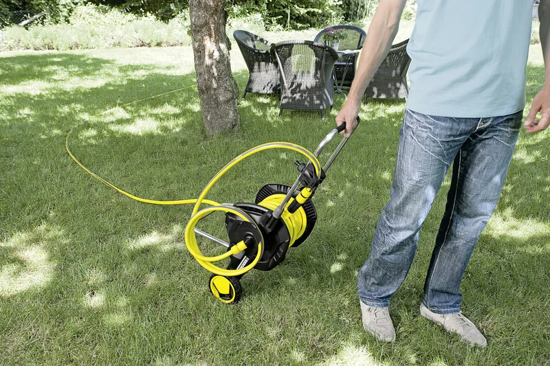 Person zieht Kärcher Schlauchwagen über Rasen. Im Hintergrund Gartenmöbel unter Baum. Outdoor-Szene bei Tageslicht.