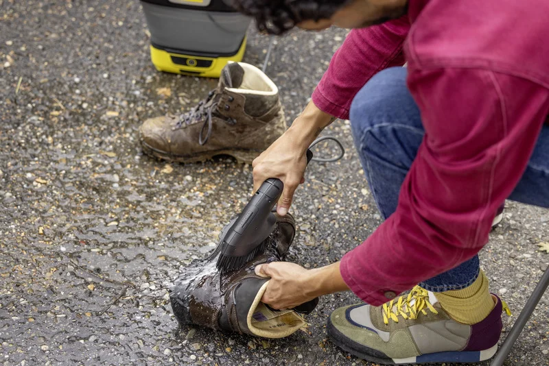 Person reinigt einen braunen Stiefel mit einem Kärcher Handgerät auf einem nassen Asphaltboden. Ein weiterer Stiefel steht daneben.