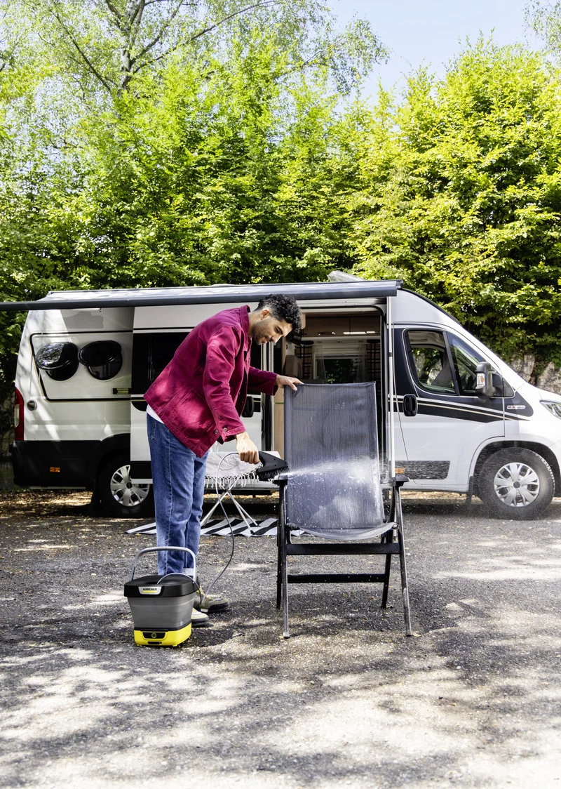 Person reinigt einen Campingstuhl mit einem Kärcher Hochdruckreiniger vor einem Wohnmobil im Freien, umgeben von grünen Bäumen.