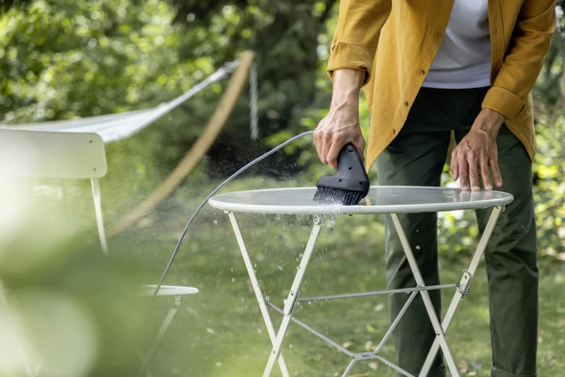 Person reinigt einen runden Gartentisch mit einer Kärcher Reinigungsbürste im Freien. Im Hintergrund sind Bäume und eine Hängematte.