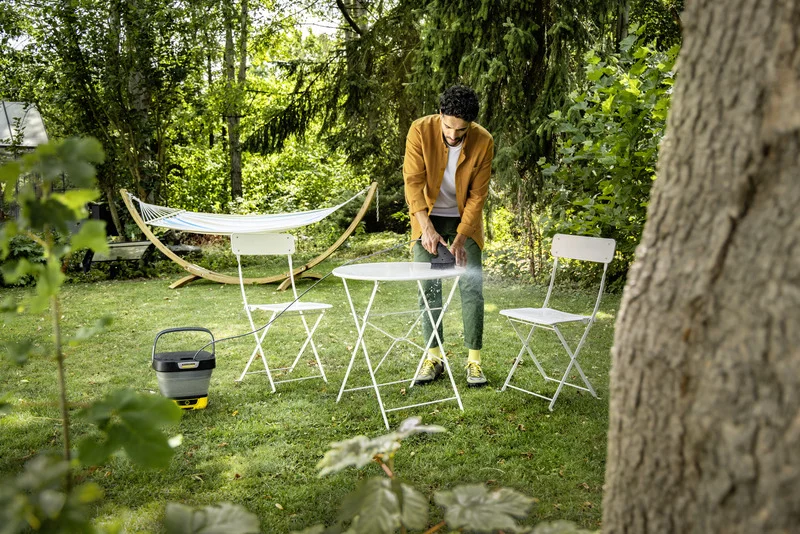 Person reinigt Tisch im Garten mit Kärcher Hochdruckreiniger, umgeben von Bäumen, Hängematte und weißen Gartenstühlen.