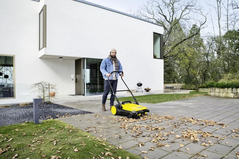Person reinigt mit Kärcher Kehrmaschine eine gepflasterte Fläche vor einem modernen weißen Gebäude, umgeben von Bäumen und Garten.