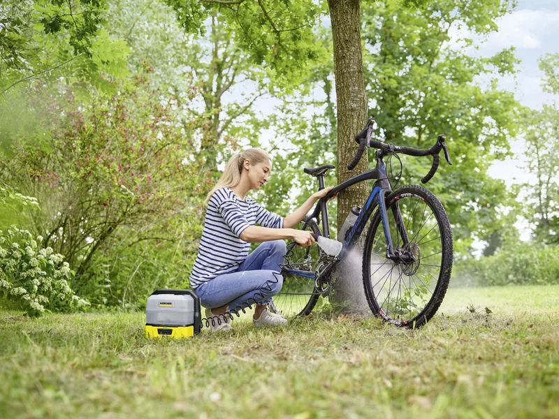 Frau reinigt Fahrrad im Freien mit Kärcher Hochdruckreiniger. Sie trägt ein gestreiftes Shirt und kniet auf einer Wiese neben einem Baum.