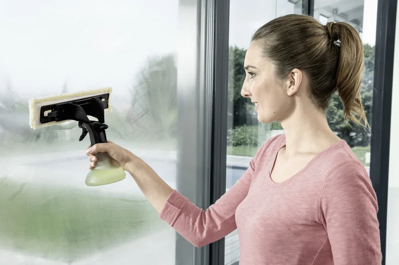 Frau reinigt Fenster mit Kärcher Fensterreiniger in einem modernen Raum, draußen ist Grünfläche sichtbar.