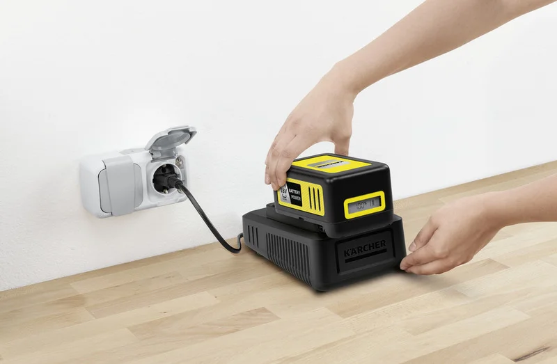 Person lädt Kärcher 18V Akku an Ladestation auf, Kabel ist in Wandsteckdose eingesteckt, Tischoberfläche aus Holz.