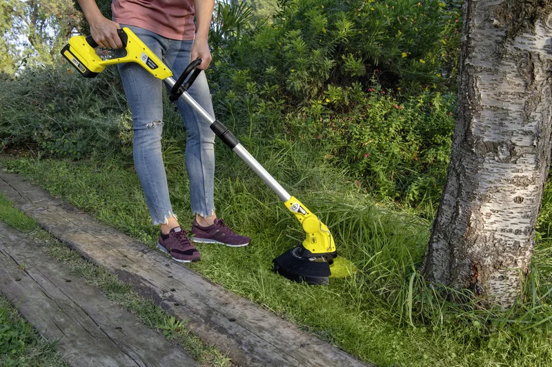Person in Jeans und Turnschuhen benutzt einen gelben Kärcher Rasentrimmer im Garten neben einem Baum.