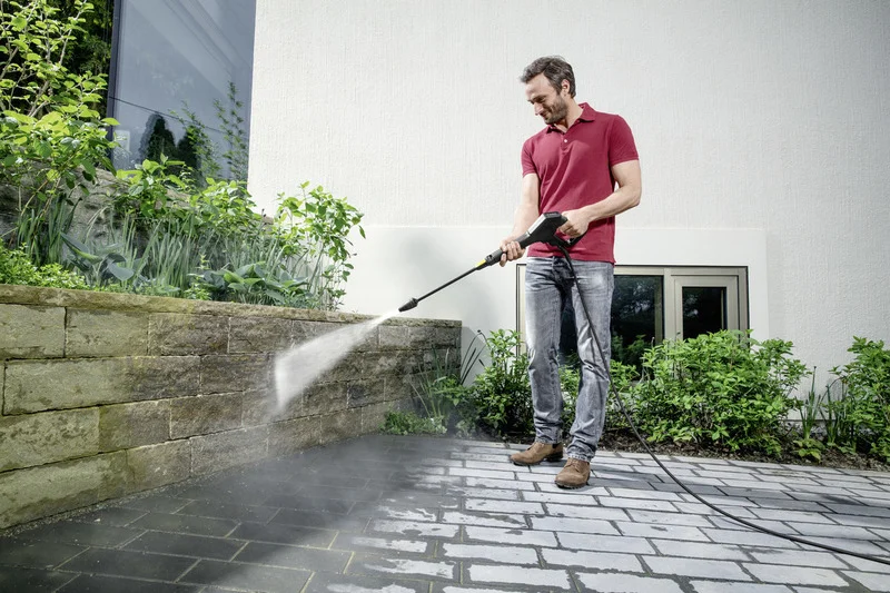Mann reinigt mit Kärcher Hochdruckreiniger eine Steinmauer im Garten. Er trägt ein rotes Polohemd und Jeans.