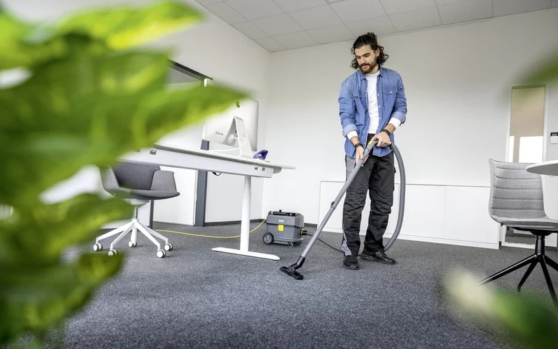 Person reinigt Büro mit Kärcher Staubsauger. Schreibtisch mit Computer, Bürostuhl und Pflanze im Vordergrund.