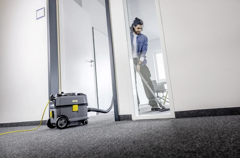 Person reinigt Teppichboden mit Kärcher Staubsauger in einem Büro. Staubsauger steht im Vordergrund, Person im Hintergrund durch Glastür sichtbar.