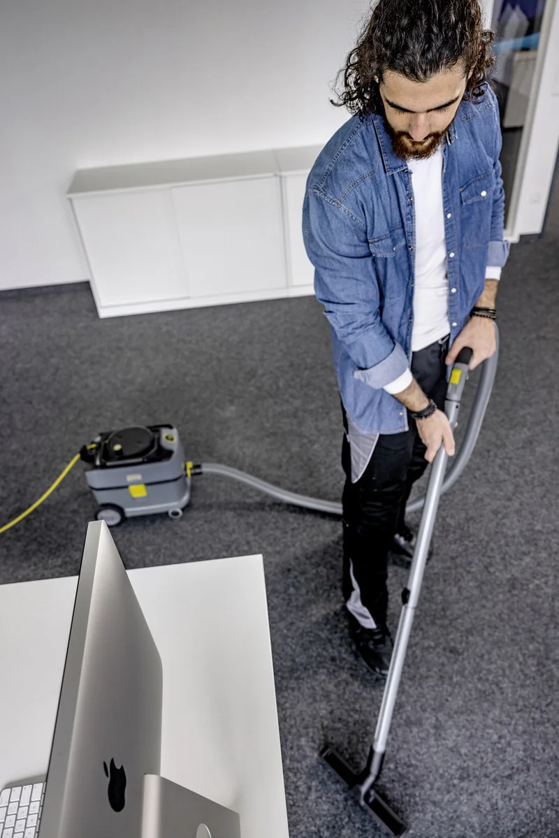 Person in Jeanshemd bedient Kärcher Teppichreiniger in Büro mit grauem Teppichboden, neben einem Computer auf weißem Tisch.