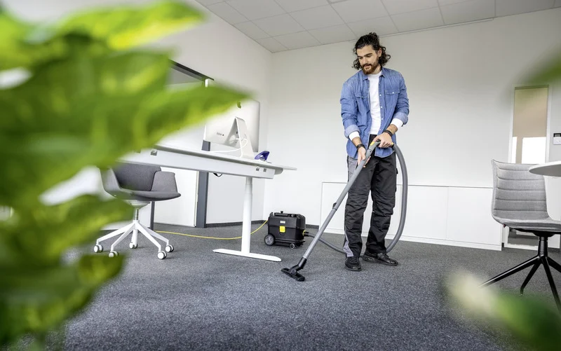 Person reinigt Büro mit Kärcher Staubsauger, umgeben von Pflanzen und Möbeln. Im Hintergrund ein Schreibtisch mit Computer.