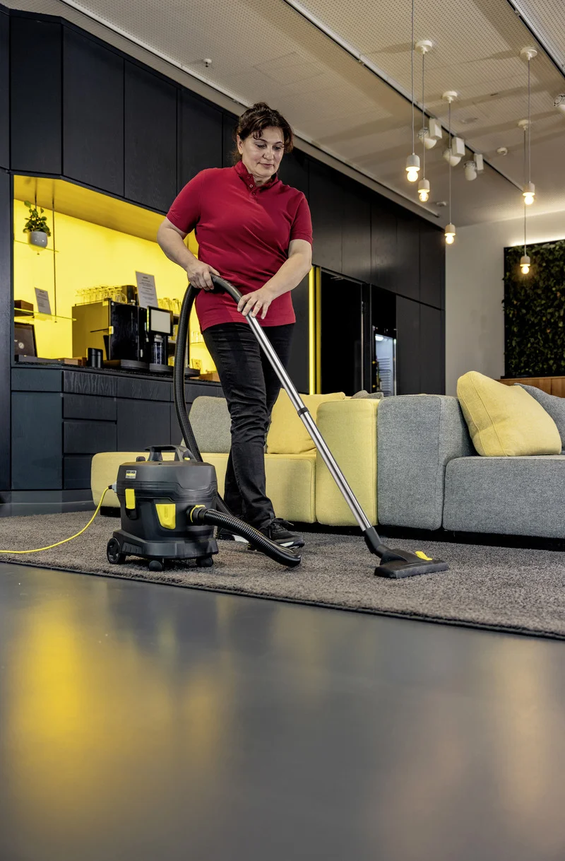 Person in rotem Shirt saugt Teppich mit Kärcher Staubsauger in modernem Wohnzimmer mit gelbem Sofa und dunkler Einrichtung.