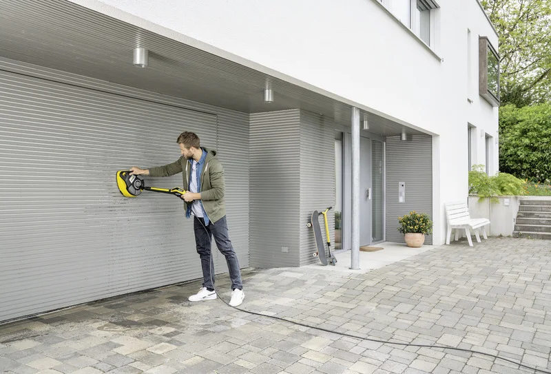 Mann reinigt Garagentor mit Kärcher Hochdruckreiniger, neben ihm ein Skateboard und ein gelber Schlauch auf einem gepflasterten Hof.
