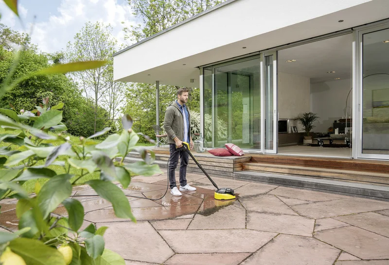 Person reinigt Terrasse mit Kärcher Hochdruckreiniger. Moderne Terrasse mit Glastüren, Pflanzen im Hintergrund.