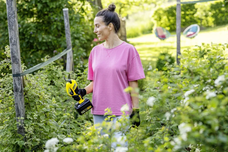 Person in rosa T-Shirt hält eine gelbe Kärcher Akku-Gartenschere in einem grünen Garten. Im Hintergrund sind zwei bunte Stühle sichtbar.