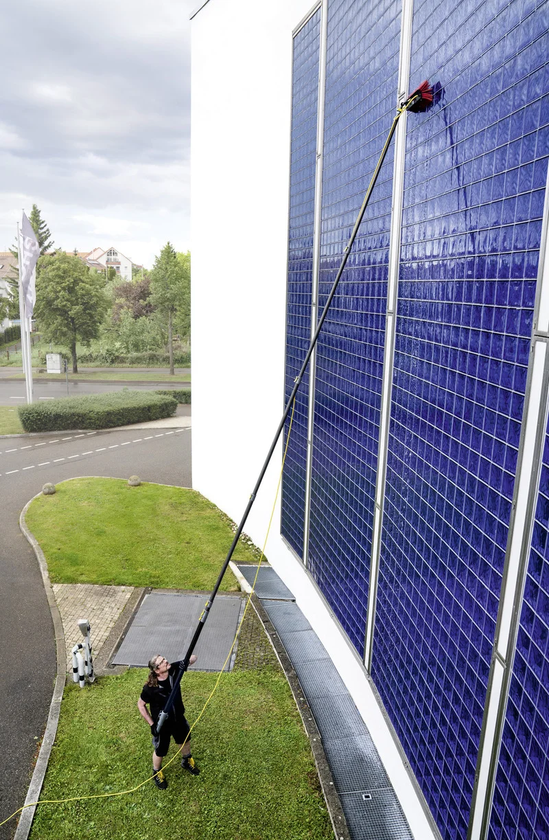 Mann reinigt mit einer langen Bürste eine große blaue Solaranlage an einem Gebäude im Freien. Wolkiger Himmel im Hintergrund.