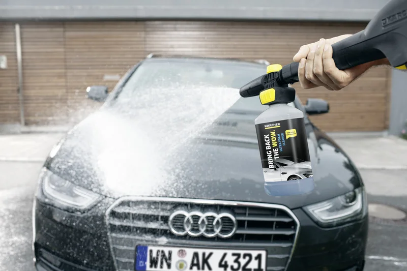 Hand hält Kärcher Schaumdüse, sprüht weißen Schaum auf schwarzen Audi, draußen vor Holzgarage.