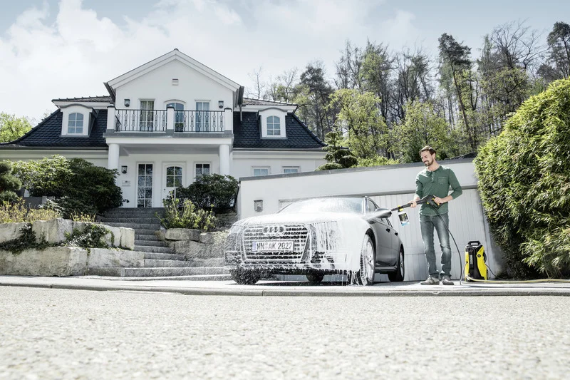 Mann reinigt Audi mit Kärcher Hochdruckreiniger vor einem großen weißen Haus, umgeben von Bäumen und Sträuchern.