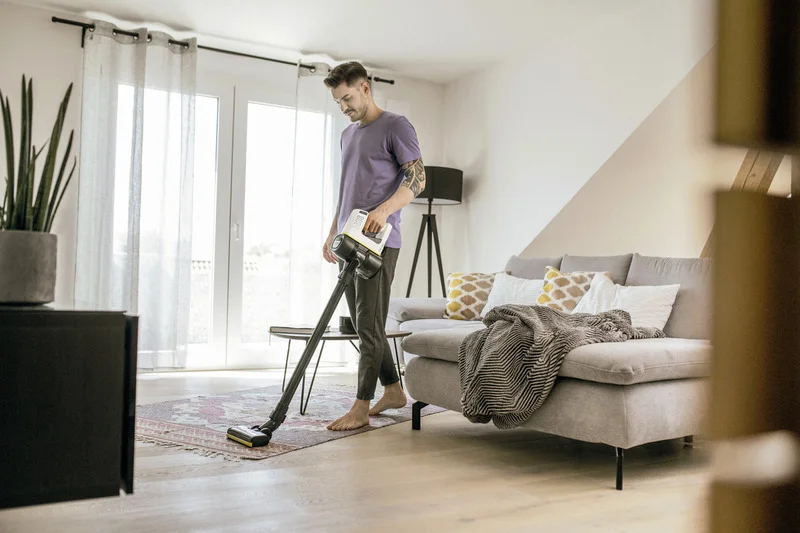 Person in lila T-Shirt benutzt Kärcher Staubsauger in modernem Wohnzimmer mit grauem Sofa und großen Fenstern.