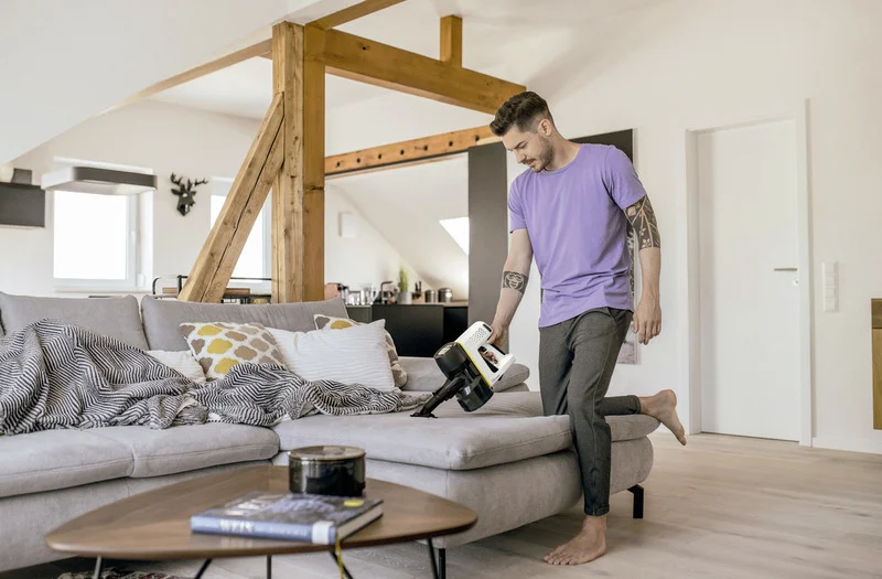 Person in lila T-Shirt und grauer Hose reinigt graues Sofa mit einem Kärcher Handstaubsauger in einem modernen Wohnzimmer mit Holzbalken.