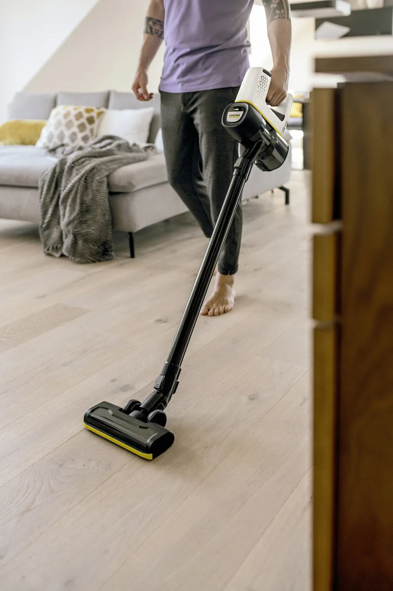 Person mit Kärcher Staubsauger auf hellem Holzboden in modernem Wohnzimmer, Sofa im Hintergrund.