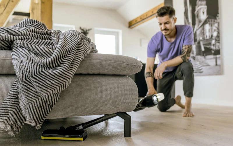 Person reinigt mit einem Kärcher Staubsauger unter einem grauen Sofa in einem hellen Wohnzimmer mit Holzbalken und Wandbild.