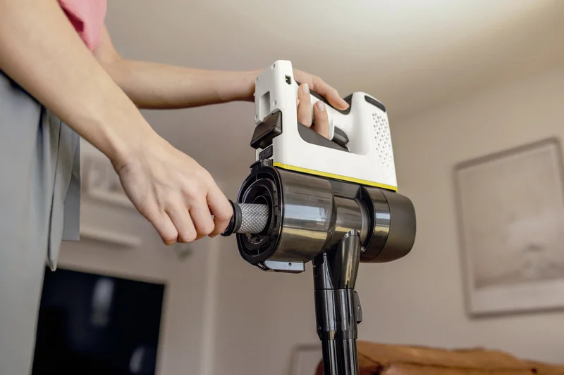 Person reinigt Filter eines Kärcher Akku-Staubsaugers in einem Wohnzimmer. Hände halten den Filter und das Gerät.
