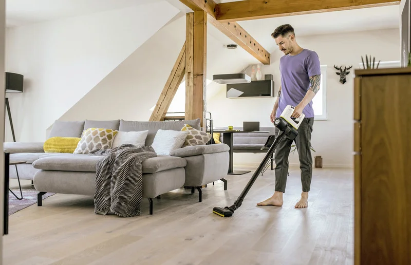 Person in lila T-Shirt saugt mit einem Kärcher Staubsauger einen hellen Holzboden in einem modernen Wohnzimmer mit grauem Sofa.