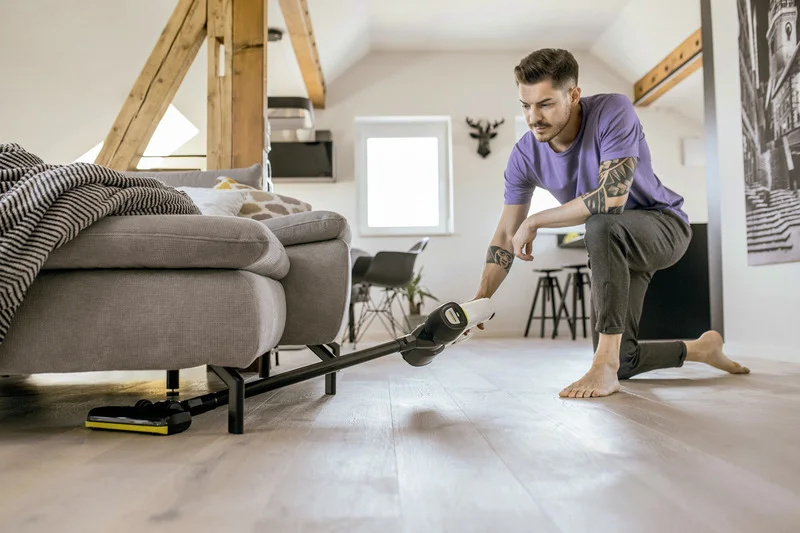 Person reinigt den Boden unter einem Sofa mit einem Kärcher Staubsauger in einem modernen Wohnzimmer mit Holzbalken.