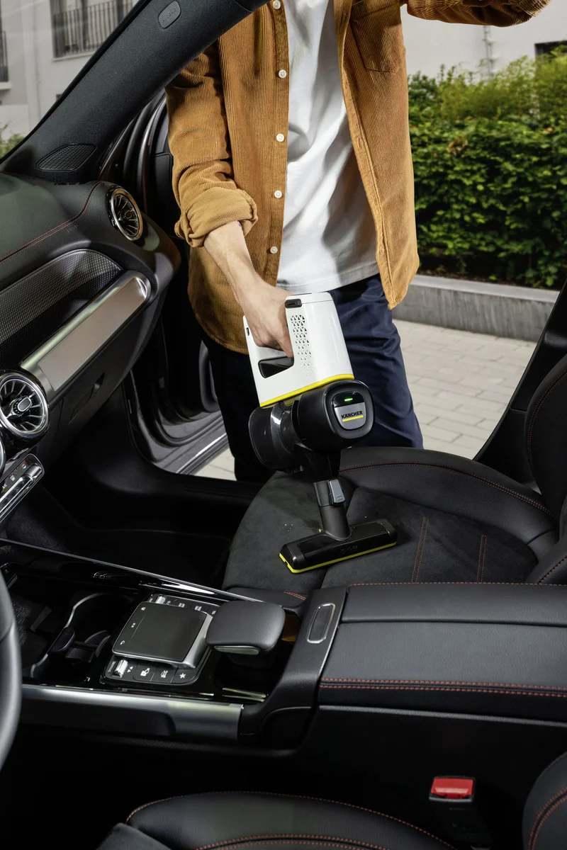 Person reinigt Autositz mit einem Kärcher Handstaubsauger. Innenraum eines Autos, braune Jacke, weißes T-Shirt.