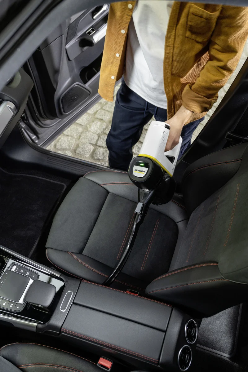 Person reinigt Autositz mit Kärcher Fenstersauger, Innenraum eines Autos, braune Jacke, schwarze Hose.