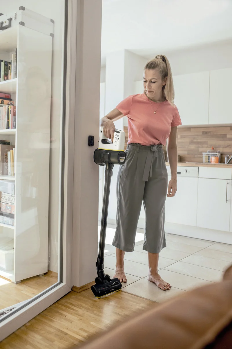 Frau in rosa Shirt und grauer Hose steht in einer modernen Küche und hält einen Kärcher Akku-Staubsauger.