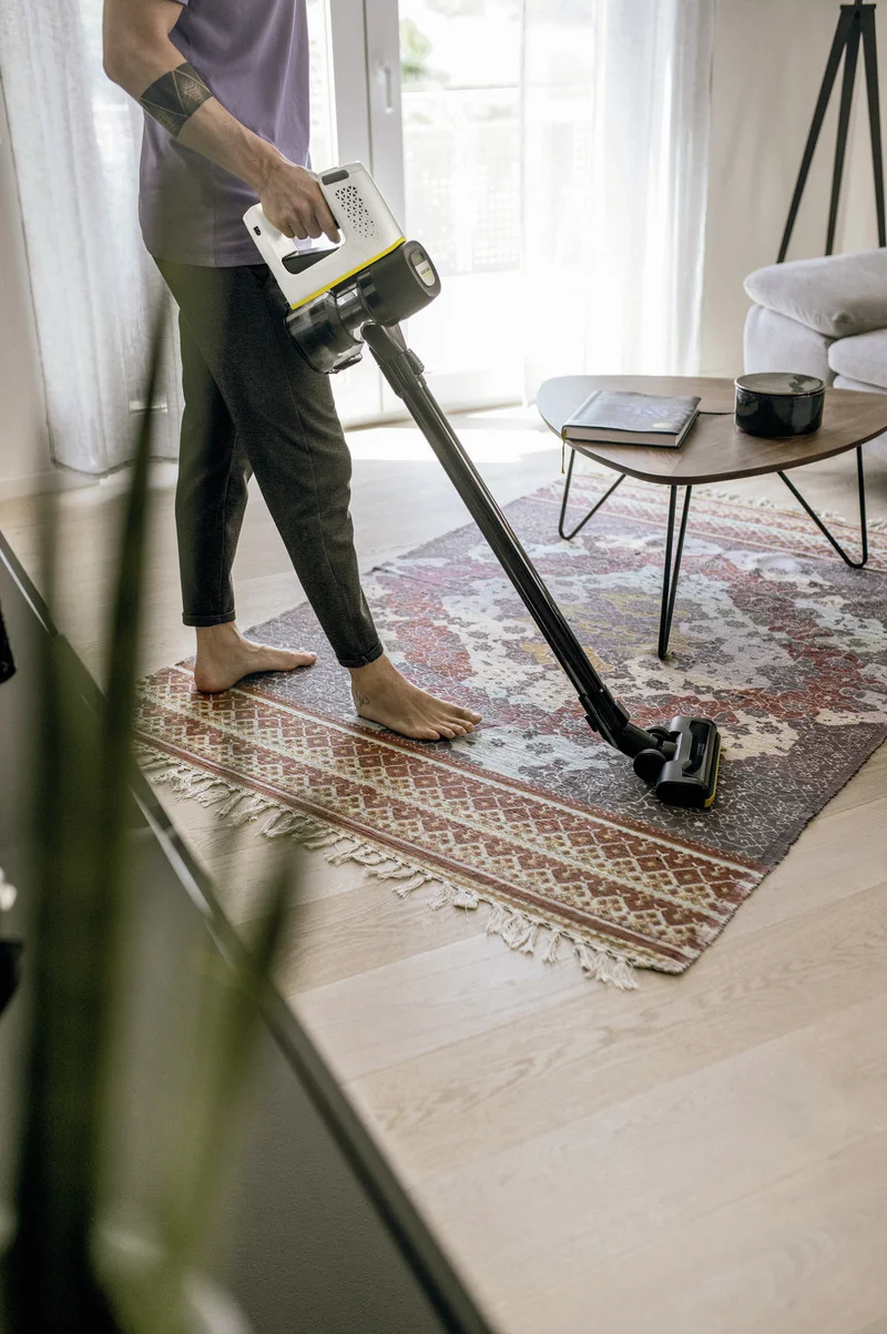 Person reinigt Teppich mit einem Kärcher Staubsauger in einem Wohnzimmer. Tisch mit Buch und Vase im Hintergrund.