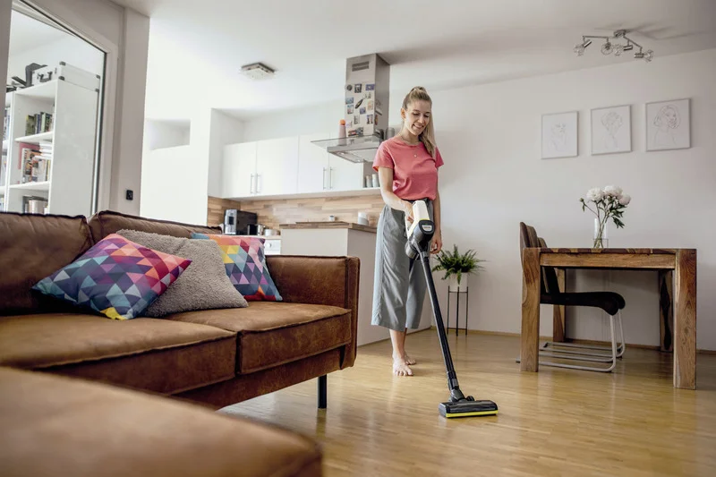 Person mit rotem Shirt und grauer Hose benutzt Kärcher Staubsauger in modernem Wohnzimmer mit braunem Sofa und Holztisch.