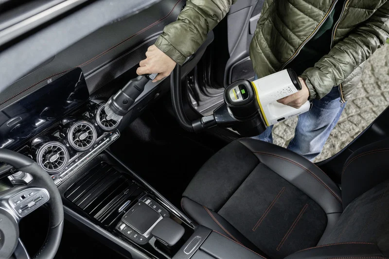 Person reinigt Autoinnenraum mit einem Kärcher Handstaubsauger, fokussiert auf Lüftungsschlitze und Mittelkonsole.