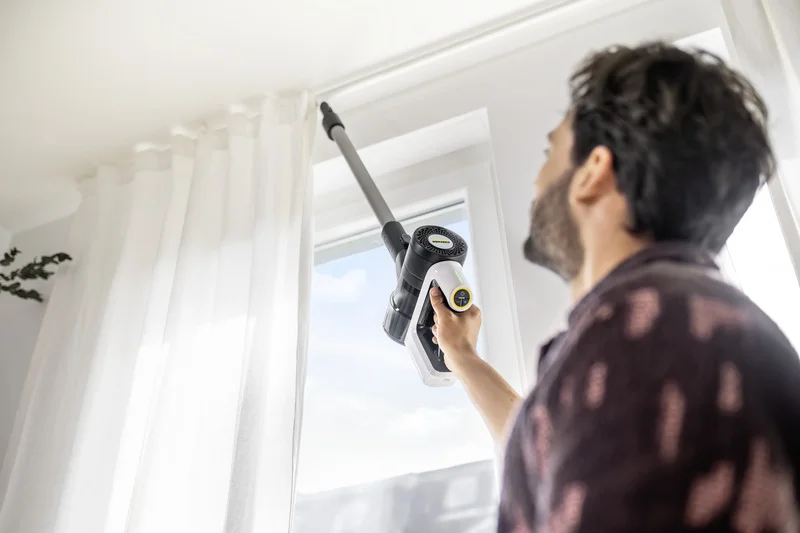 Person reinigt Fensterrahmen mit einem Kärcher Handstaubsauger in einem hellen Raum mit weißen Vorhängen.